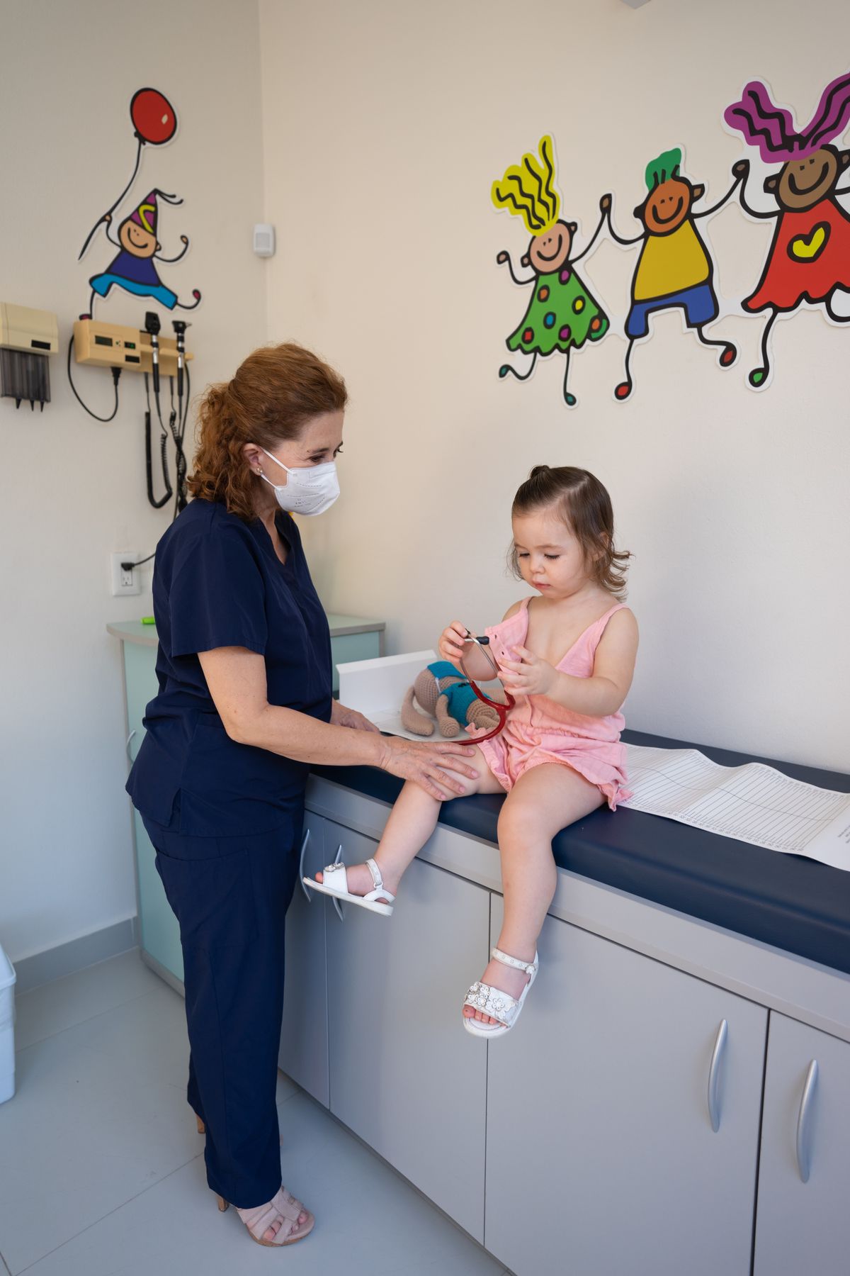 A nurse examining a toddler sitting on an examination couch in a decorated clinic room.